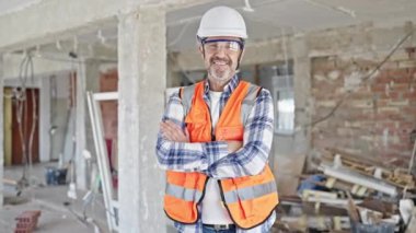 Orta yaşlı inşaatçı kendinden emin gülümsüyor inşaat alanında kollarını kavuşturmuş duruyor.