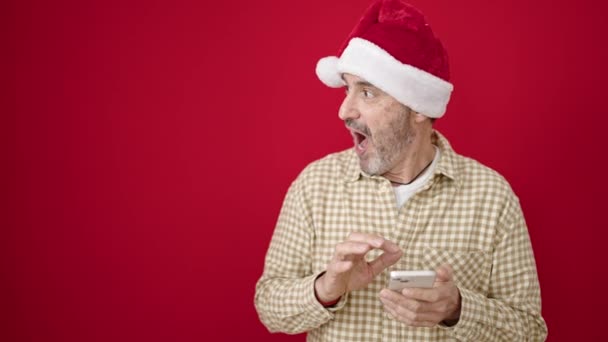 中年男身に着けているクリスマス帽子使用スマートフォン上の隔離された赤の背景 — ストック動画