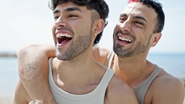 İki erkek, deniz kenarında birbirlerine sarılarak gülümsüyor.