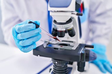 Young beautiful hispanic woman scientist using microscope pouring blood on sample at laboratory