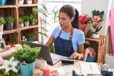 Young beautiful hispanic woman florist using laptop and calculator at florist