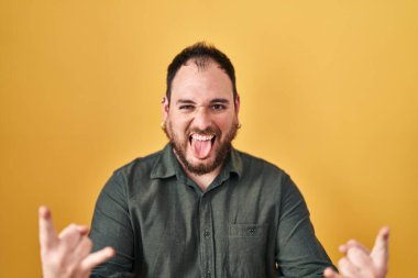 Plus size hispanic man with beard standing over yellow background shouting with crazy expression doing rock symbol with hands up. music star. heavy music concept. 
