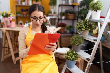 Young arab woman florist writing on document sitting on chair at flower shop