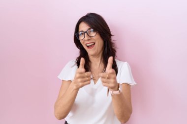 Orta yaşlı, beyaz tişörtlü ve gözlüklü İspanyol bir kadın kamerayı mutlu ve komik yüzüyle işaret ediyor. iyi enerji ve titreşimler. 