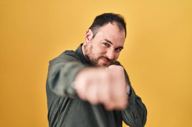 Plus size hispanic man with beard standing over yellow background punching fist to fight, aggressive and angry attack, threat and violence 