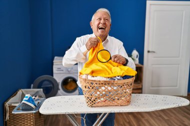 Senior man with grey hair holding magnifying glass looking for stain at clothes smiling and laughing hard out loud because funny crazy joke. 