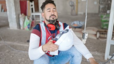 İnşaat alanında el fanı olarak kasketi kullanan genç Latin bir inşaatçı yerde oturuyor.