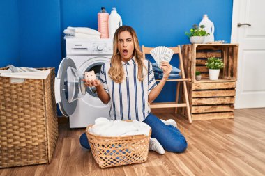Young blonde woman doing laundry saving money in shock face, looking skeptical and sarcastic, surprised with open mouth 
