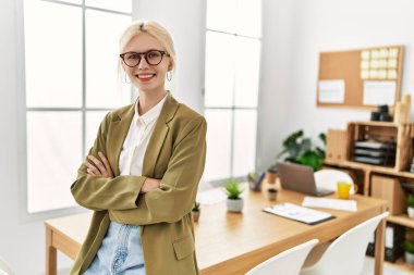Genç, sarışın, iş kadını, gülümseyen, kendine güvenen, kollarını kavuşturup ofiste el kol kola duran.