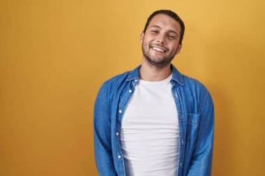 Sarı arka planda dikilen İspanyol adam ellerini kavuşturup gülümseyerek gevşemiş ve neşeliydi. Başarı ve iyimserlik 