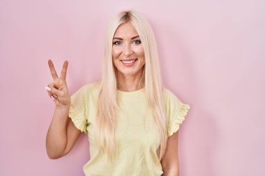 Pembe arka planda duran beyaz kadın iki numarayı gösterip işaret ederken kendinden emin ve mutlu bir şekilde gülümsüyor.. 