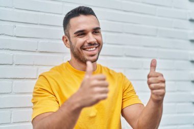 Young hispanic man smiling confident doing ok sign with thumbs up over isolated white background