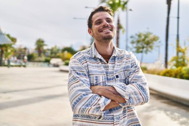 Genç İspanyol adam gülümsüyor. Parkta kollarını kavuşturmuş duruyor.
