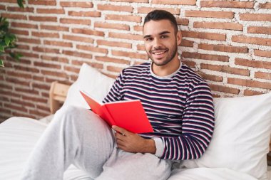 Young hispanic man reading book sitting on bed at bedroom