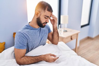 Young hispanic man taking pills for headache sitting on bed at bedroom