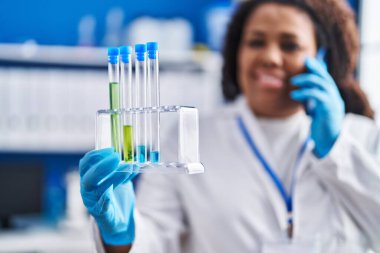 Afrika kökenli Amerikalı bilim kadını laboratuvarda elinde test tüpleriyle akıllı telefondan konuşuyor.