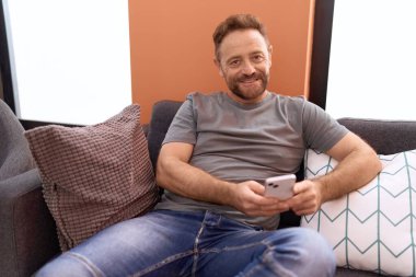 Middle age man using smartphone sitting on sofa at home