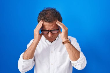 Orta yaşlı İspanyol bir adam mavi arka planda, elleri başının üstünde duruyor. Stres yüzünden baş ağrısı çekiyor. migren ağrısı. 