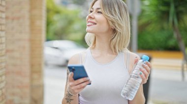 Genç sarışın kadın sokakta elinde su şişesi tutan akıllı telefon kullanıyor.