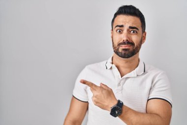 Beyaz arka plan üzerine gündelik giysiler giymiş, sakallı genç İspanyol bir adam endişeli ve endişeli bir ifade takınıyor. 
