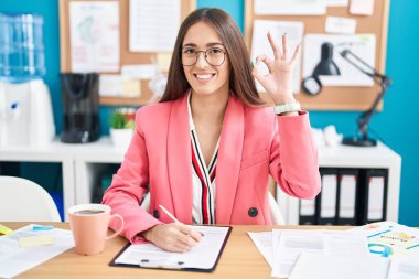 Young hispanic woman working at the office wearing glasses smiling positive doing ok sign with hand and fingers. successful expression. 