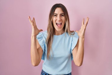 Young hispanic woman standing over pink background celebrating mad and crazy for success with arms raised and closed eyes screaming excited. winner concept 