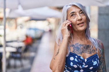 Orta yaşlı, kır saçlı, gülümseyen, kendine güvenen bir kadın caddedeki akıllı telefondan konuşuyor.