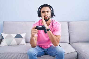 Hispanic young man playing video game holding controller sitting on the sofa hand on mouth telling secret rumor, whispering malicious talk conversation 