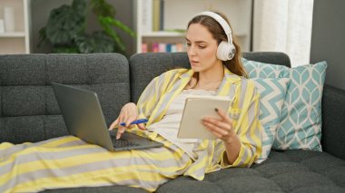 Evde dizüstü bilgisayar ve kulaklık kullanan genç sarışın kadın.