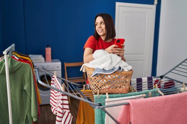Young beautiful hispanic woman using smartphone hanging clothes on clothesline at laundry room