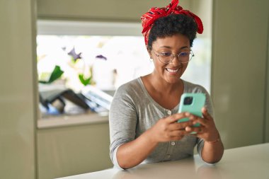 Afro-Amerikalı kadın akıllı telefon kullanıyor. Evde masada oturuyor.