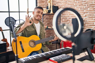Young hispanic man doing online guitar tutorial doing ok sign with fingers, smiling friendly gesturing excellent symbol 