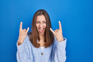 Mavi arka planda duran genç bir kadın çılgın bir ifadeyle el kaldırarak rock sembolü yapıyor. Müzik yıldızı. ağır müzik konsepti. 
