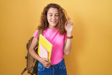 Young caucasian woman wearing student backpack and holding books gesturing finger crossed smiling with hope and eyes closed. luck and superstitious concept. 