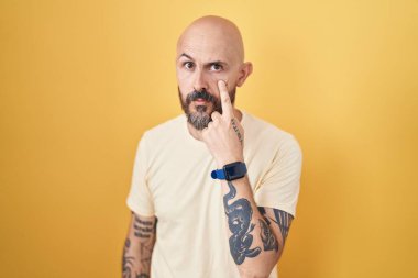 Hispanic man with tattoos standing over yellow background pointing to the eye watching you gesture, suspicious expression 