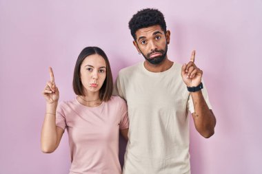 Pembe arka planda genç İspanyol çift üzgün ve üzgün görünüyor, parmaklarıyla yön gösteriyor, mutsuz ve depresif.. 