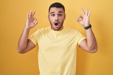 Young hispanic man standing over yellow background looking surprised and shocked doing ok approval symbol with fingers. crazy expression 