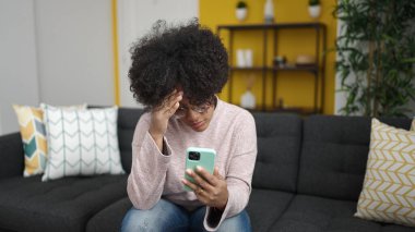 Akıllı telefon kullanan Afrikalı genç bir Amerikalı kadın evdeki kanepede otururken stresli bir şekilde oturuyor.
