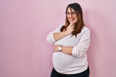 Pembe arka planda duran hamile kadın kameraya kendinden emin bir şekilde bakıyor kollarını kavuşturmuş ve çenesini kaldırmış gülümsüyor. Olumlu düşünmek. 