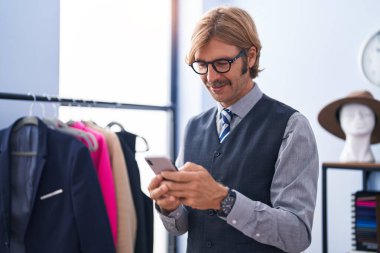 Elbise fabrikasında akıllı telefon kullanan kendine güvenen genç sarışın terzi.