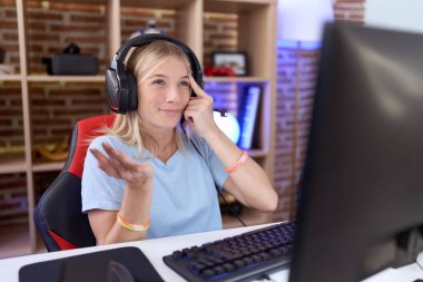 Video oyunları oynayan, kulaklık takan, kafası karışmış ve avuç içi açık bir şekilde fotokopi çeken ve alnını işaret eden beyaz bir kadın. Bunu bir düşün.. 