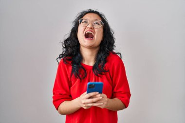Genç Asyalı kadın akıllı telefon kullanıyor kızgın ve kızgın bağırıyor öfkeyle bağırıyor, öfkeyle yukarı bakıyor. 