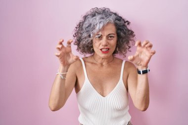 Pembe arka planda gri saçlı orta yaşlı bir kadın komik bir şekilde gülümsüyor kedi gibi pençe hareketi yapıyor, agresif ve seksi bir ifade. 