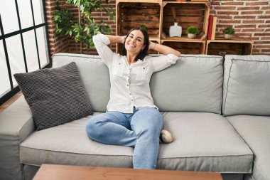 Genç, güzel, İspanyol bir kadın. Elleri başının üstünde, evdeki kanepede oturuyor.