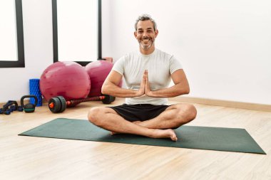 Orta yaşlı, gri saçlı, gülümseyen, spor merkezinde yoga eğitimi alan bir adam.