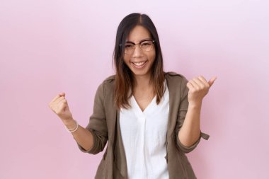 Orta yaşlı Çinli bir kadın pembe arka planda gözlük takıyor. Çok mutlu ve heyecanlı. Kolları havada, gülümseyen ve başarı için çığlık atan kazanan bir jest yapıyor. kutlama konsepti. 