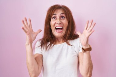 Middle age woman standing over pink background celebrating mad and crazy for success with arms raised and closed eyes screaming excited. winner concept 
