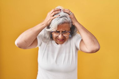 Middle age woman with grey hair standing over yellow background suffering from headache desperate and stressed because pain and migraine. hands on head. 