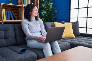 Evdeki kanepede dizüstü bilgisayar kullanan genç Latin kadın.