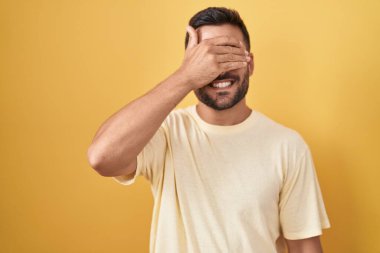 Sarı arka planda dikilen yakışıklı İspanyol adam. Gülümsüyor ve gülümsüyor. Gözlerini şaşkınlıkla kapatıyor. kör kavram. 